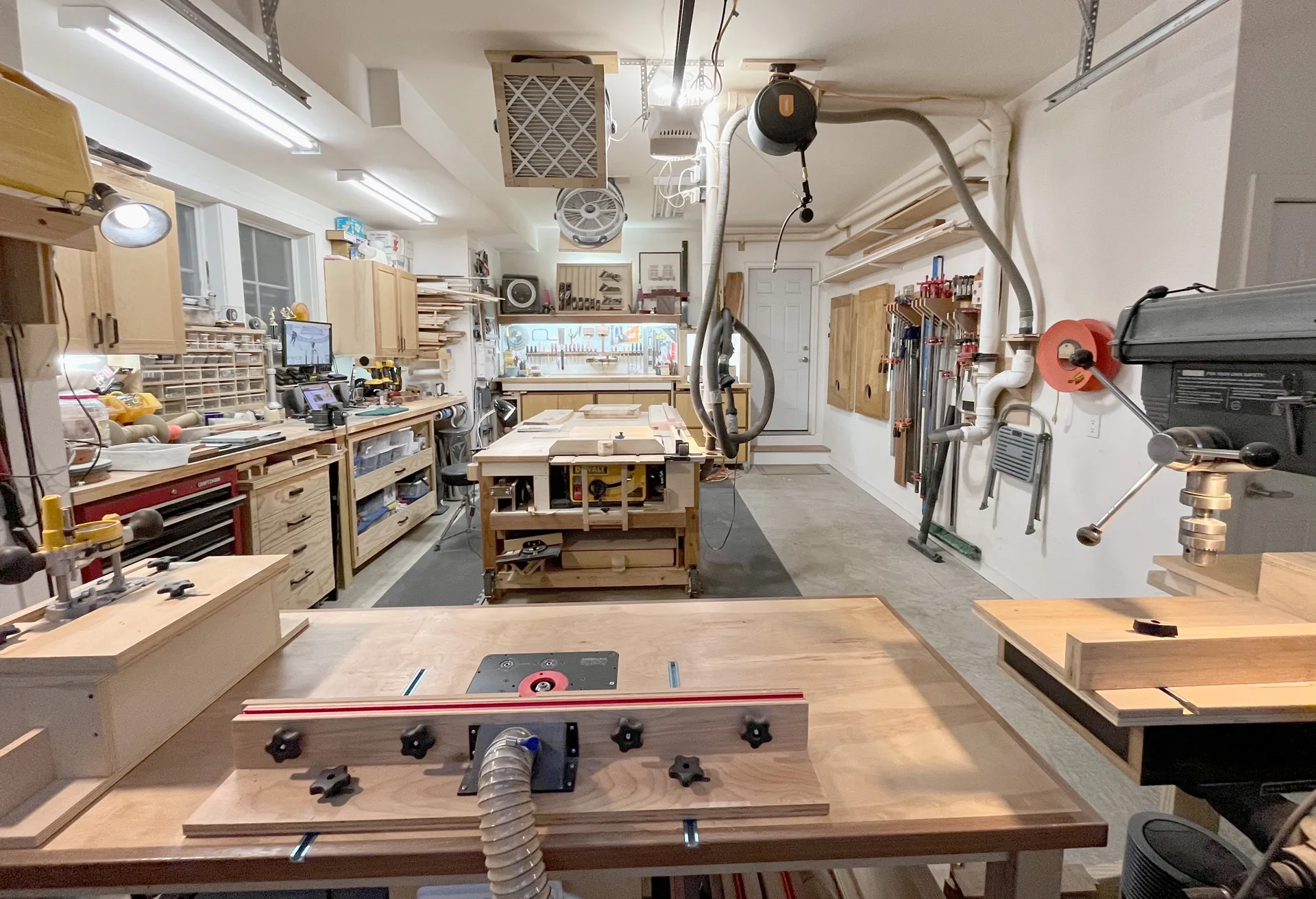 Wide view of the garage woodworking studio, with workbenches, a table saw, router table, drill press, dust-collection hoses, and wall-mounted clamp storage