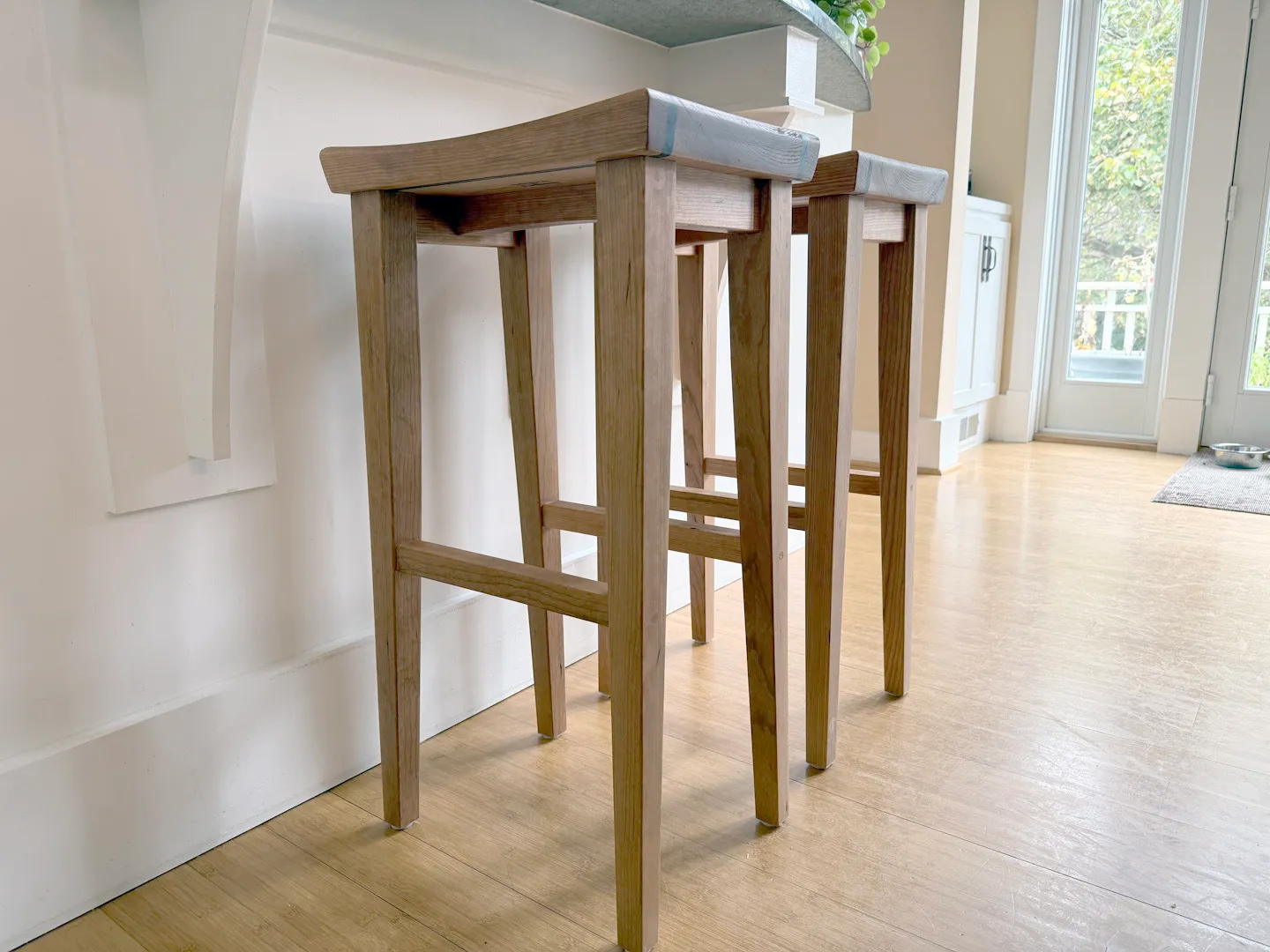 Two finished cherry bar stools with saddle seats under a kitchen island