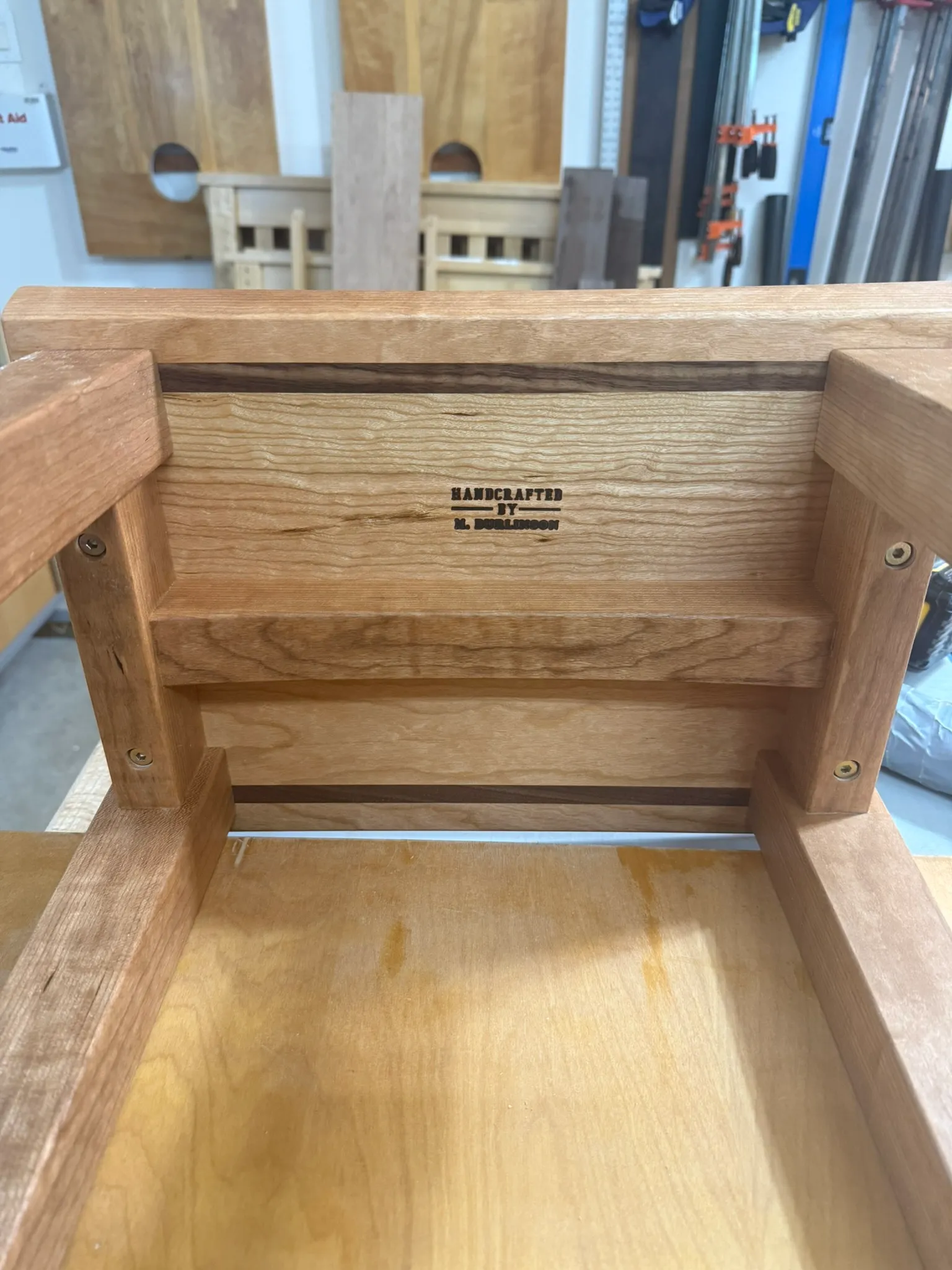 Underside of a cherry bar stool seat, showing the "Handcrafted by M. Burlinson" branded stamp and a walnut inlay stripe